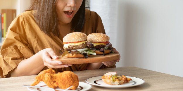 hungry-overweight-woman-holding-hamburger-wooden-plate-fried-chicken-pizza-table_41689-829